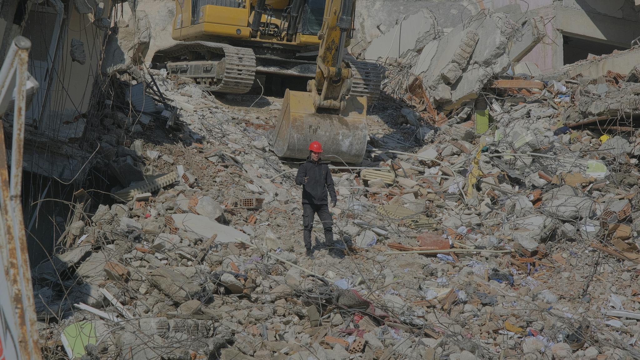 Excavator clearing urban demolition site rubble with a worker on site.